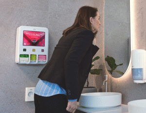 Dameshygiëne met menstruatiedispenser van Tork met Libresse tampons, inlegkruisjes en maandverband. Vouw kijkt in spiegel op zakelijk toilet